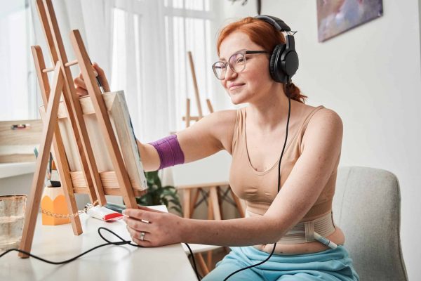 Indoor image of creative professional female artist with renal failure listening music, looking at her picture and having inspired facial expression while painting in her living room
